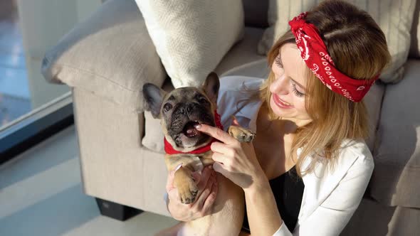 Young Woman in a White Shirt and Jeans Sits on the Floor Near the Sofa with Her Small French Bulldog