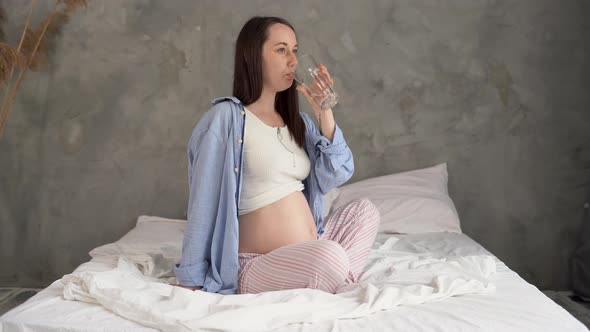 Lifestyle Portrait of a Young Pregnant Woman Sitting on the Bed and Drinking Water From a Glass