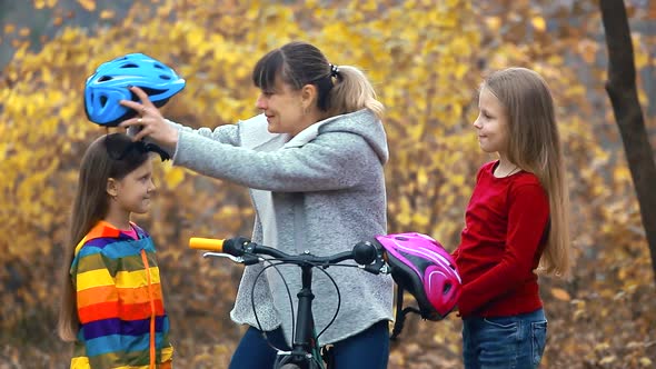 Mom helps two daughters put on a safe helmet before riding a bike on autumn day in nature