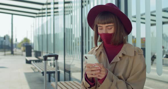 Woman in Cotton Prottective Mask Using Mobile Phone While Sitting on Bench Near Railway Station