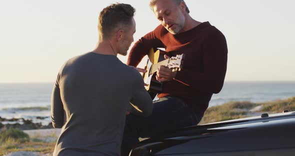 Happy caucasian gay male couple sitting on car playing guitar and talking at the beach