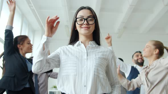 Beautiful Asian Lady Touching Glasses and Smiling Standing at Corporate Party with People Dancing
