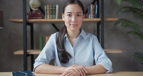 Smiling Young Asian Woman Teacher Sitting at the Table and Looking at Camera