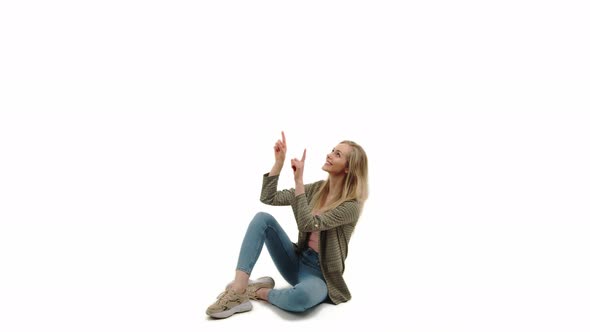 Pleasant Caucasian Blonde Girl Sitting on the Floor and with a Finger Pointing Upwards Full Shot