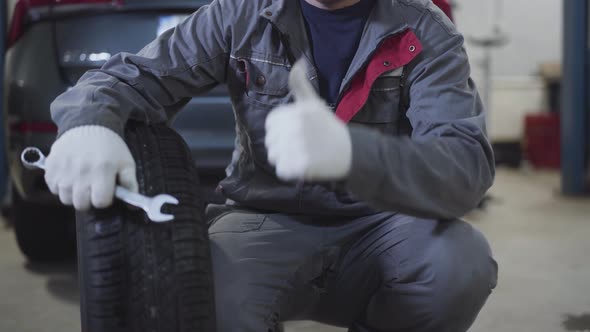 Unrecognizable Caucasian Man in Workwear Sitting with Car Wheel and Wrench and Showing Thumb Up