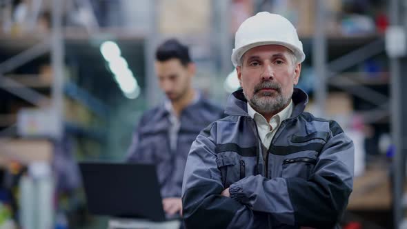 Portrait of Serious Confident Senior Man in Hard Hat Crossing Hands Looking at Camera As Blurred