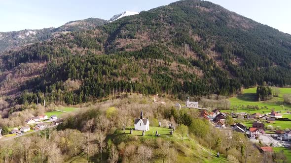 Drone Shot (facing forwards,ing forwards) of a Chapel on top of a Hill in front of Alpine Mountains