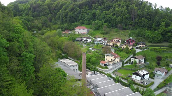 Drone going up in front of small factory chimney with settlement between green nature in back.