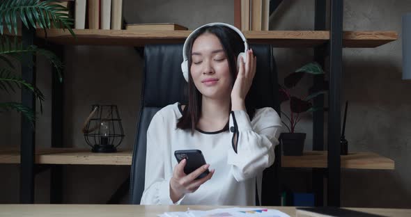 Young Asian Woman Sitting at the Table Holding Smartphone Listening to Music with Headphones in