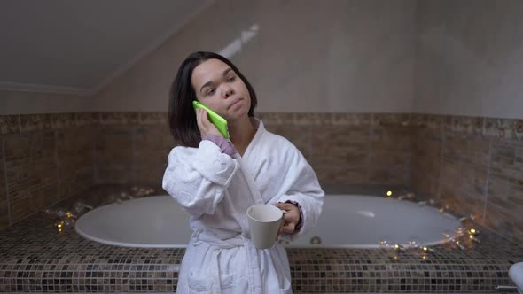 Confident Young Little Woman Talking on Phone Drinking Coffee Standing in Bathroom at Home Indoors