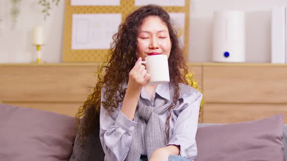 Asian peaceful woman holding cup enjoy favorite tea or coffee morning drink while sitting at sofa in