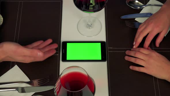 A Man and a Woman Sit at a Table, a Smartphone with a Green Screen, They Talk About Something