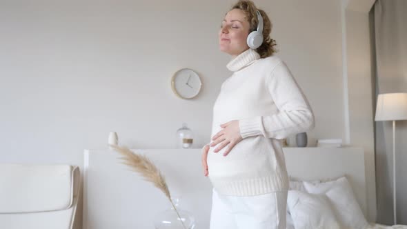 Young Pregnant Woman Wearing Headphones And Cozy Sweater Dancing At Home