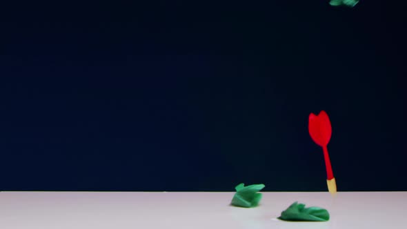 Red Dart Blows Up Green Balloon Lying on White Table Against Blue Background