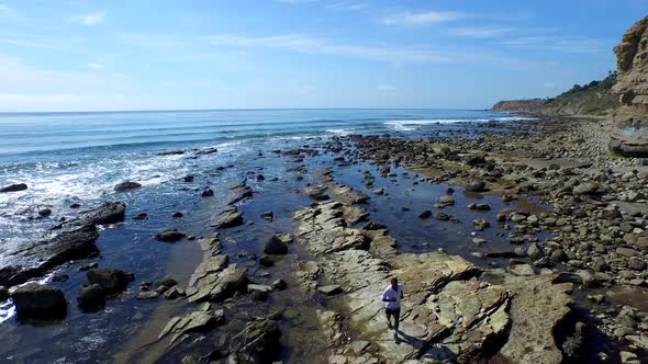 Tracking shot of a young man running on a rocky ocean beach shoreline.