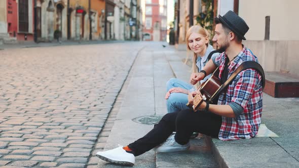 Mman and Woman Sitting on Sidewalk, Playing Guitar and Having Rest