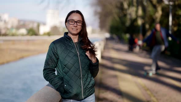 Charming Middleaged Woman in Jacket is Standing on River Embankment in Small Town