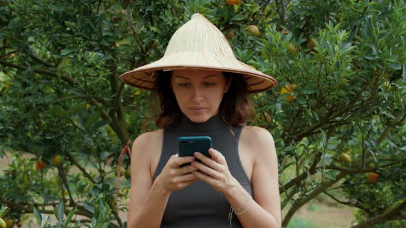 Woman Wearing a Vietnamese Hat Using Her Phone in an Orange Orchard Looking Straight at the Camera