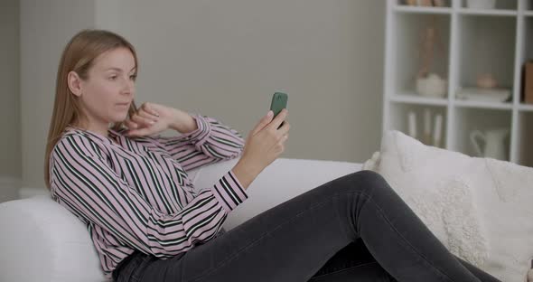 Adult Woman Is Browsing Social Nets in Smartphone, Lying on Couch at Home, Swiping and Scrolling