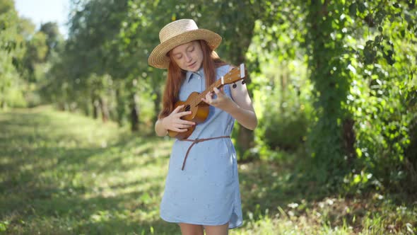 Medium Shot Redhead Teenage Girl in Blue Dress and Straw Hat Playing Ukulele Standing in Sunshine in