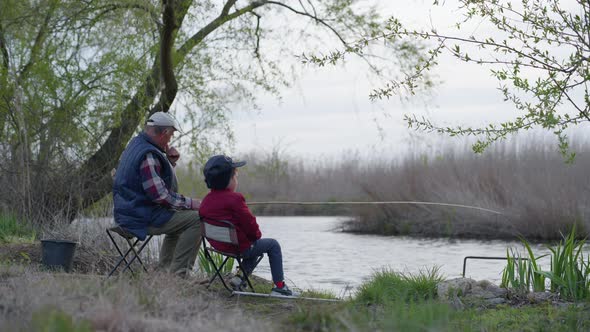 Fishermen, an Elderly Man and His Grandson in Hats Are Sitting Near the Shore and Fishing Small Fish