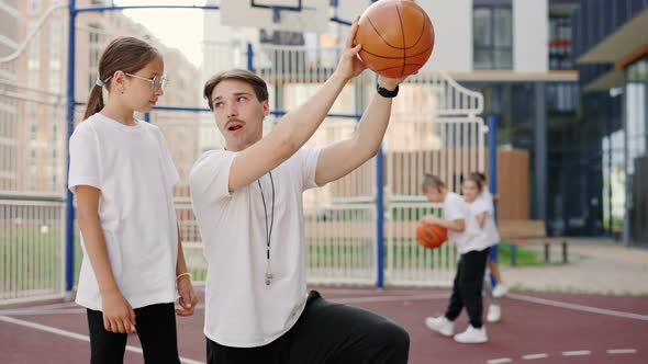 Handsome Male Coach Explaining to Child How to Put a Ball Into the Basket on Court Near School