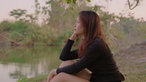 Thai Young Woman Sits Pensively By Lake in Park at Sunset