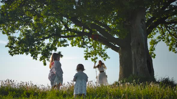 Two Daughters Walking Along Green Field To Their Mother, Swinging on a Rope Swing Tied To Old Tree.