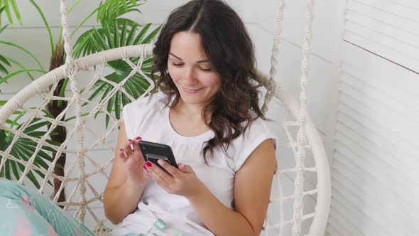 Attractive Brunette Talking on the Phone While Sitting on a Swing at Home. Woman in Home Clothes