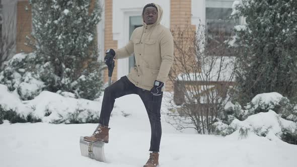 Wide Shot Portrait of Thoughtful Young African American Man Standing with Shovel Outdoors on Snowy