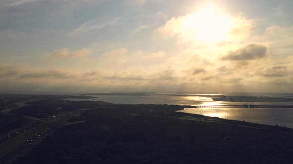 aerial view over the marsh & road facing the golden sunset as a bird investigates the drone in fligh