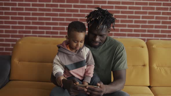 Portrait of African American Father Sitting with Son on Sofa Watching Cartoon on Smartphone