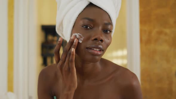 Portrait of smiling african american woman with towel using cream on her face in bathroom