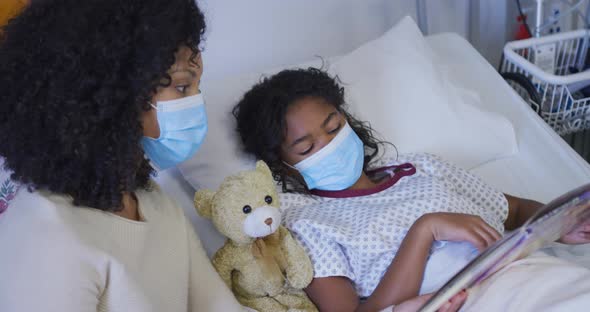 Mixed race mother and daughter lying on hospital bed reading book, both wearing face masks