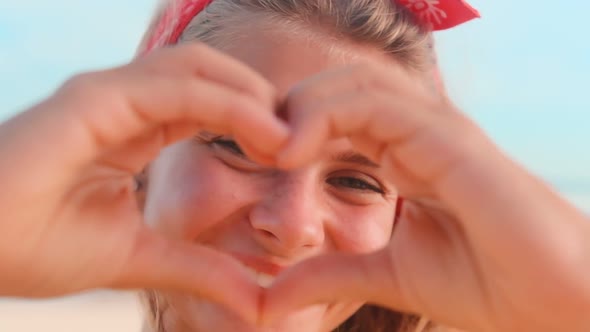 Young Cheerful Caucasian Woman Shows Gesture of Heart Stands on Beach By Ocean