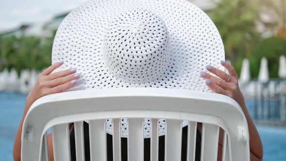 Fashionable Female Sunbathing in White Elegance Hat Relaxing on Deck Chair Near Swimming Pool