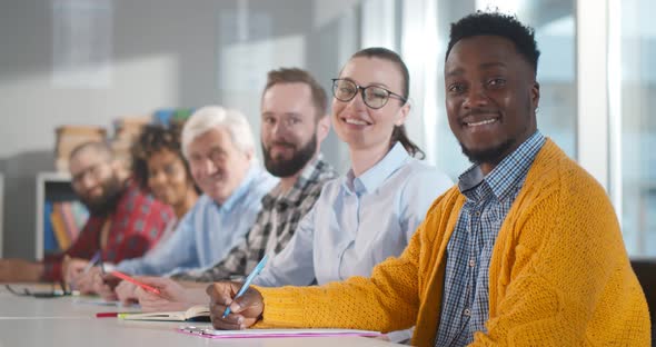 Multiethnic Adult People Sitting in Row at Desk and Smiling at Camera in Classroom