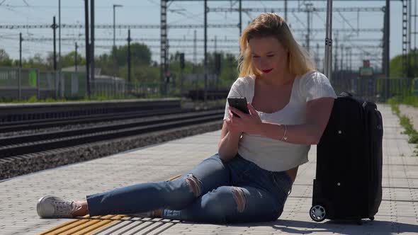 A Young Woman Sits By a Suitcase on a Train Station Platform and Works on a Smartphone, Then Smiles
