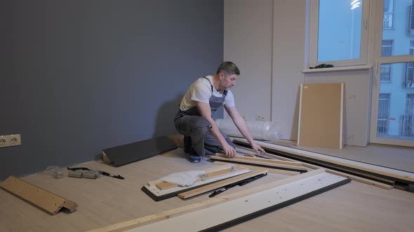 A Builder in a Work Overalls is Assembling a Bed in a New Apartment