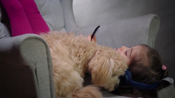 Adorable Girl Is Playing with a Small Puppy and Listens To Music on Chair