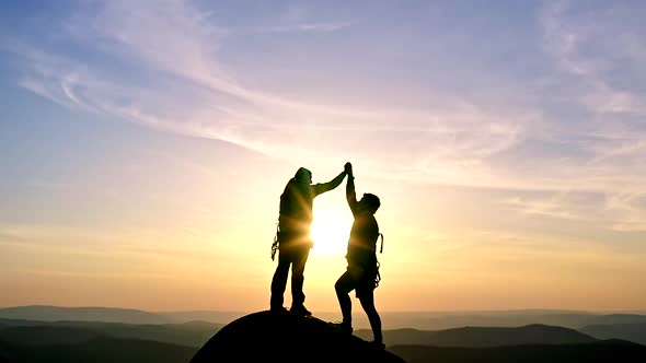 Silhouettes of a Man and a Woman Giving High Fives on Top of a Rocky Mountain Range at Sunset
