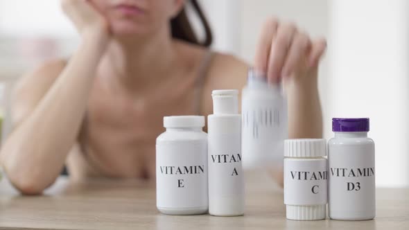 Blurred Thoughtful Woman Sitting at Table with Row of Vitamin Bottle Choosing Pills