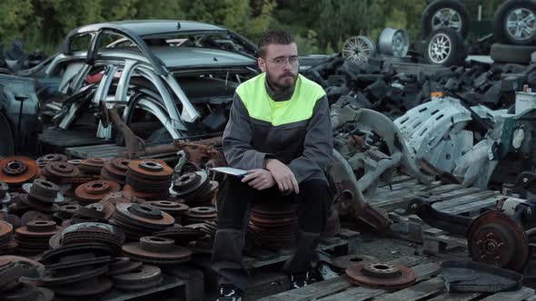 Tired Young Bearded Mechanic with Glasses Sits on a Pile of Old Disks Among Pallets with Spare Parts