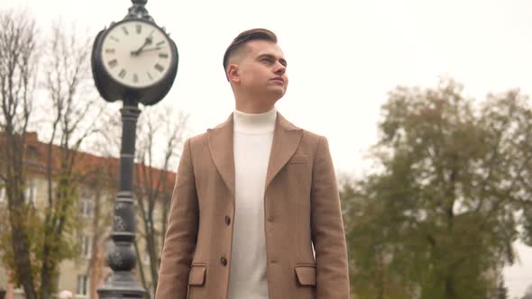 Portrait of a Handsome Fashionable Man in Brown Coat Against the Background of Street Clocks. New