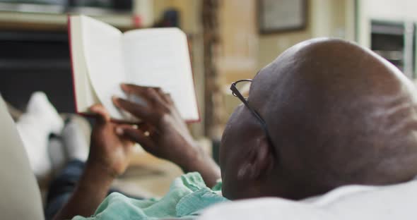 Over shoulder view of african american senior man relaxing, lying on couch reading a book