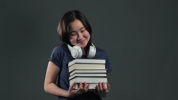 Asian Student Girl with Headphones on Grey Background Holds Stack of Books