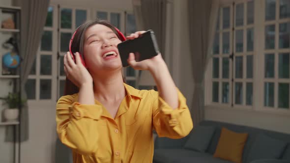 Funny Young Asian Lady Hold Sing Telephone, Wear Headphones In Living Room