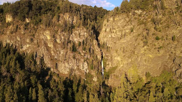 Aerial flying over Corbata Blanca thin waterfall between pine tree forest mountains at golden hour,