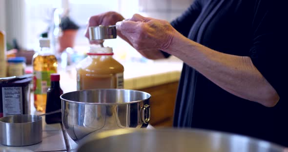 An elderly woman measuring ingredients and pouring them into a metal mixing bowl while baking a vega