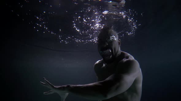 an African Man with a Naked Torso Floats in the Dark Water. He Moves His Hands. Bubbles Float on the